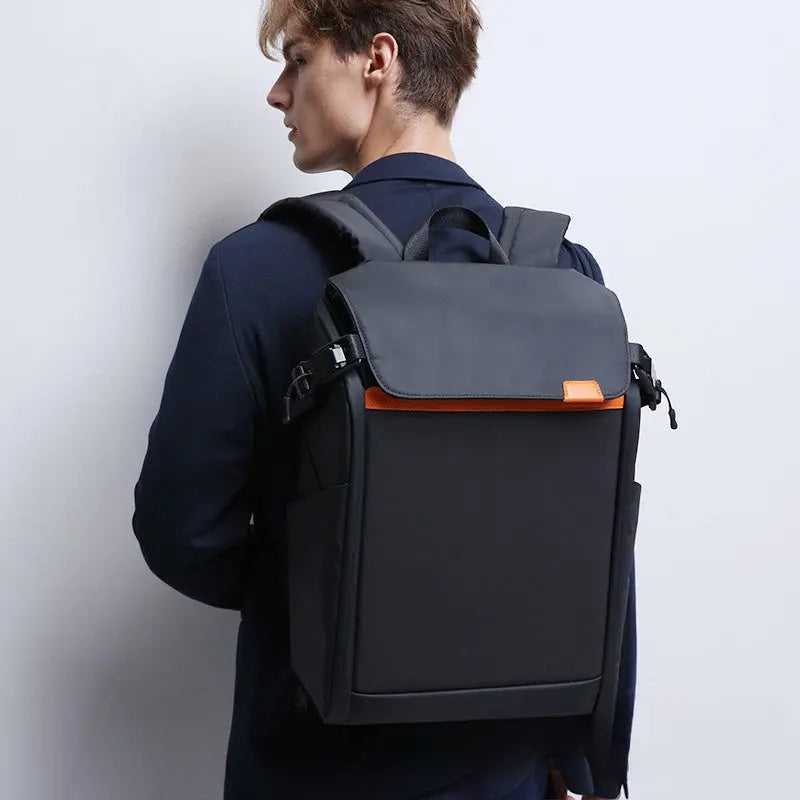 A person in a dark blazer carries the Dropshipman Bluetooth Projection Backpack—black, water-resistant, with orange trim—viewed from the back and side against a plain light-colored wall.