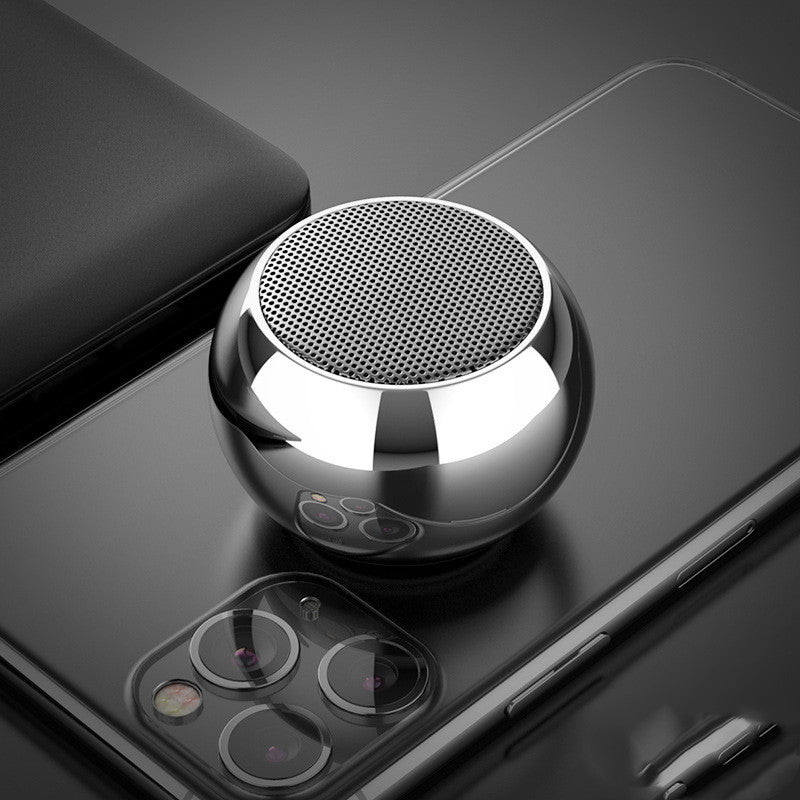 A compact, silver Dropshipman M3 Mini Colorful Wireless Speaker sits atop a modern triple-camera smartphone, both arranged on a sleek, dark surface.