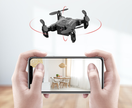 A person holds a smartphone showing a bright dining room, while the Dropshipman Mini folding drone with red indicator lights hovers indoors, highlighting its remote control quadcopter features.
