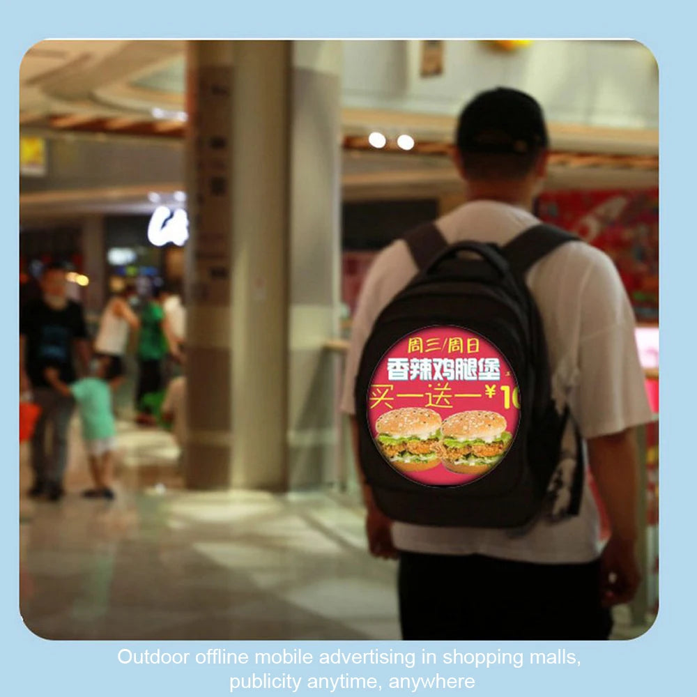 A person in a shopping mall dons a HeadphoneHeat BSIDE 3D Holographic Projector Backpack, showcasing an advertisement on its back. The ad, featuring Chinese text, promotes a buy-one-get-one-free deal on burgers with vivid images and the price prominently displayed on a high-resolution display. The caption reads, 