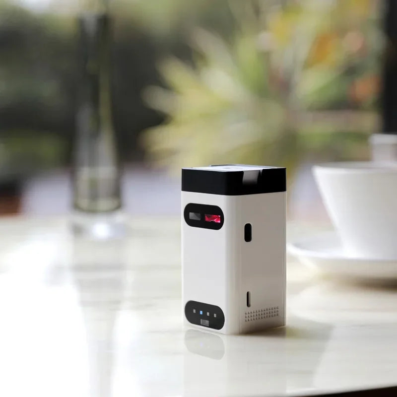 A compact white Dropshipman Bluetooth Virtual Laser Keyboard projector sits on a marble table, with a blurred background showing a vase, cup, and greenery.
