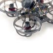Close-up of the HappyModel Mobula6 2024 1S 65mm Ultra Light Micro FPV Drone2 by HeadphoneHeat, featuring a SuperX Flight Controller with protective circular guards around its propellers. The drone's camera and electronic components are visible, showcasing a sleek black and gray design with advanced ELRS 2.4GHz technology for seamless control.