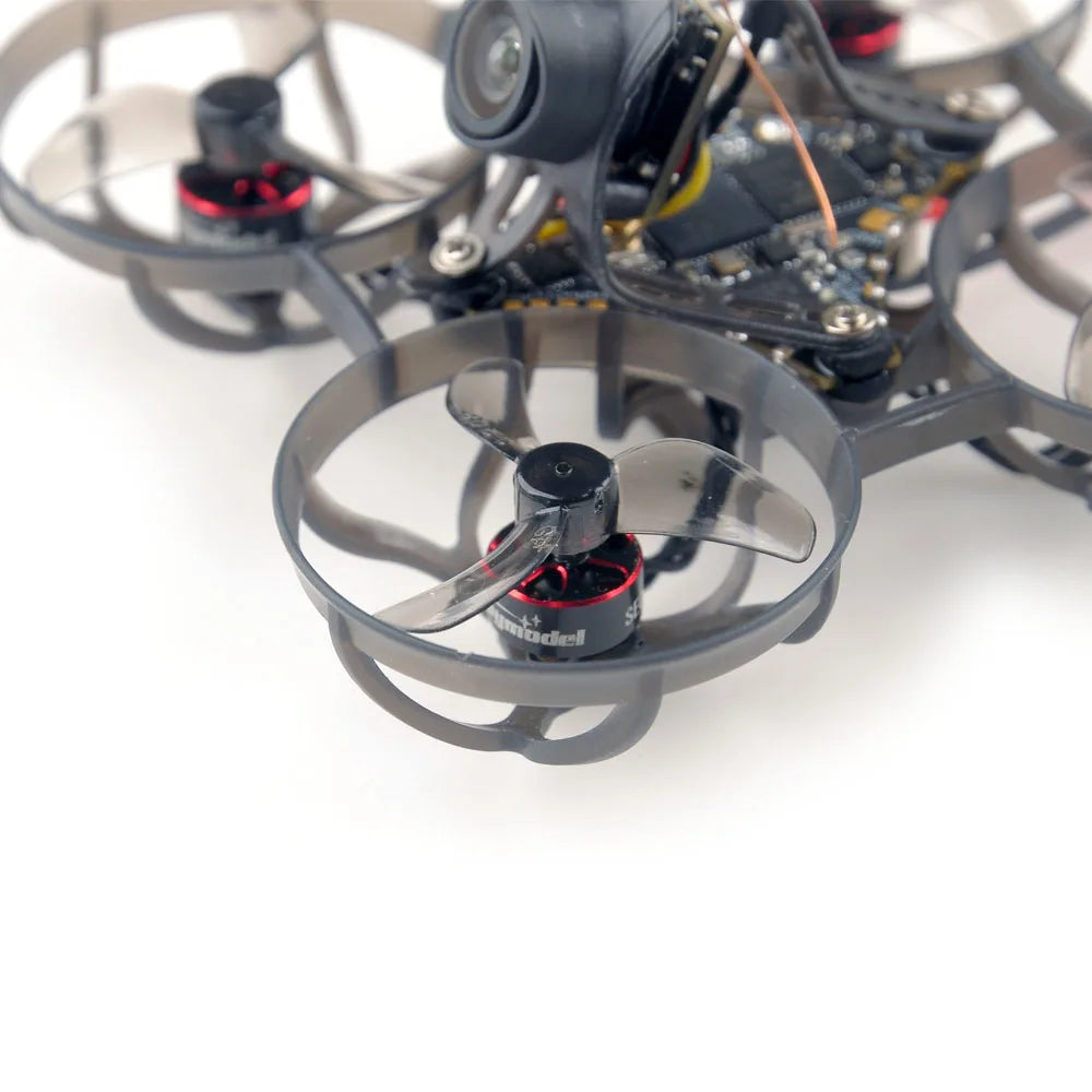 Close-up of the HappyModel Mobula6 2024 1S 65mm Ultra Light Micro FPV Drone2 by HeadphoneHeat, featuring a SuperX Flight Controller with protective circular guards around its propellers. The drone's camera and electronic components are visible, showcasing a sleek black and gray design with advanced ELRS 2.4GHz technology for seamless control.