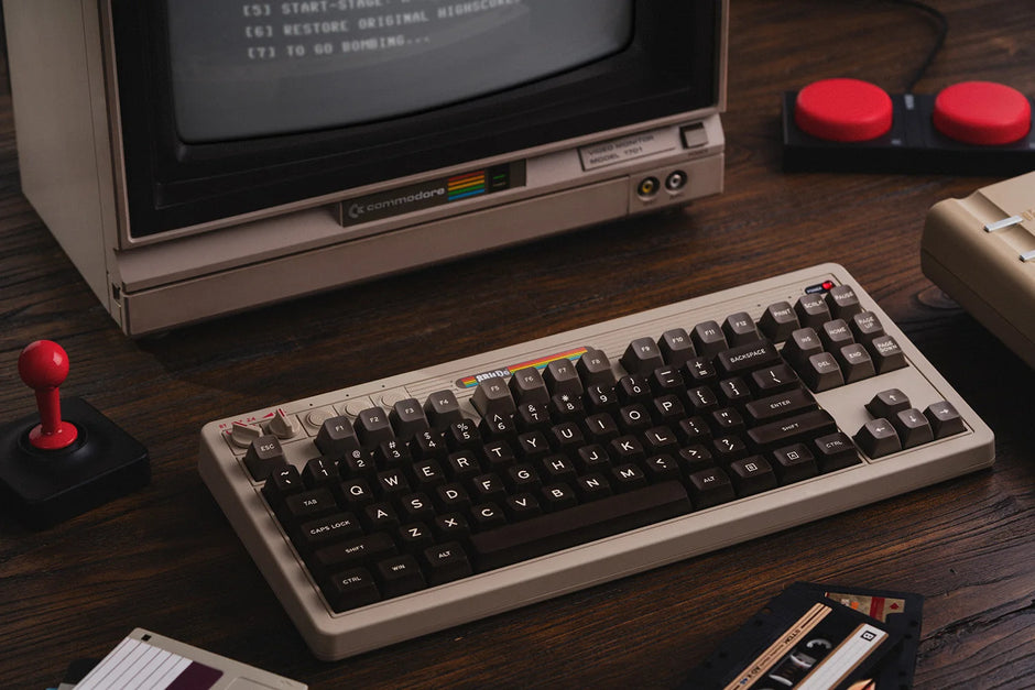 A vintage computer setup includes an 8BitDo Retro Mechanical Keyboard by HeadphoneHeat and a classic Commodore monitor on a wooden desk. Nearby, you'll find a joystick, a floppy disk, a cassette tape, and two red-button controllers. The screen shows a blurred command prompt.