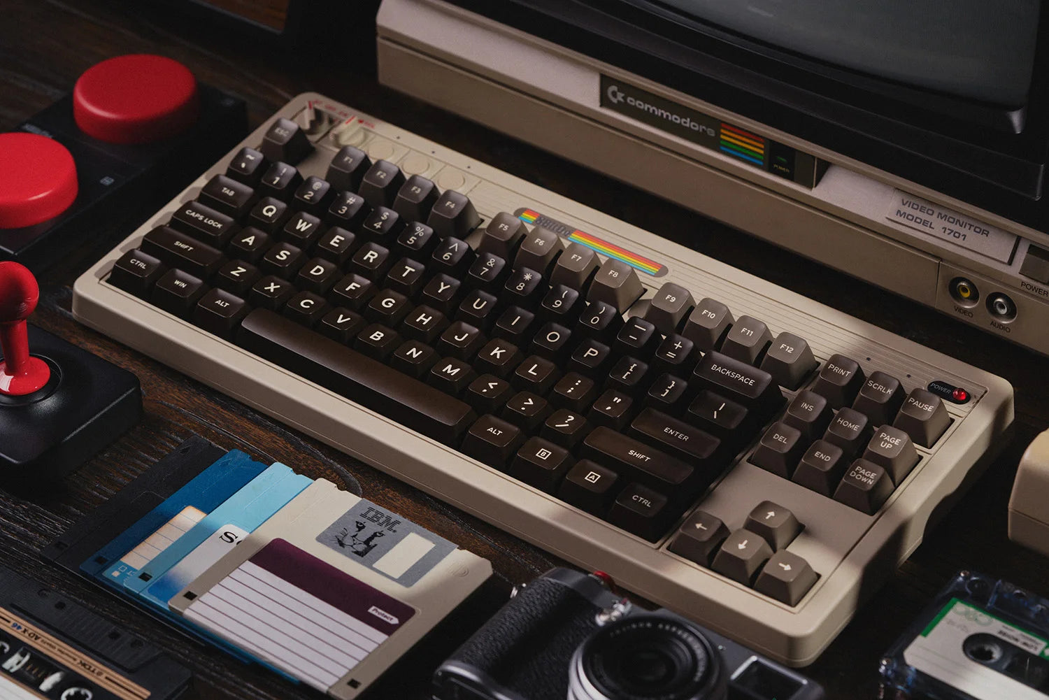 A vintage computer setup featuring a classic Commodore computer with a brown and beige keyboard, floppy disks, a joystick, and a camera creates a retro technology vibe. Nostalgic gaming and computing accessories are enhanced by the 8BitDo Retro Mechanical Keyboard from HeadphoneHeat, which perfectly ties the aesthetic together.