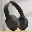 A pair of HeadphoneHeat V5.3 Over-Ear Headphones with a lightweight fit in sleek black rests upright on a light wooden surface against a white wall, softly lit. Enjoy wireless Bluetooth connectivity and high-quality audio.