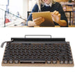 A person is smiling while using a tablet in the background. In the foreground, there's a HeadphoneHeat Retro Typewriter Mechanical Keyboard with round keys, wooden base, and Bluetooth 5.0 connectivity, resembling a classic typewriter.