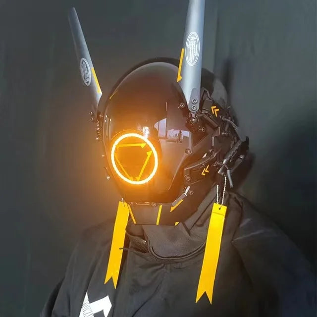 A person wearing the HeadphoneHeat LED Lights Cyberpunk Full Face Mask featuring glowing orange accents, long vertical antennae, and dangling yellow tags. The visor showcases an illuminated circular design enhanced by LEDs, with a dark background highlighting the mask's striking glow.