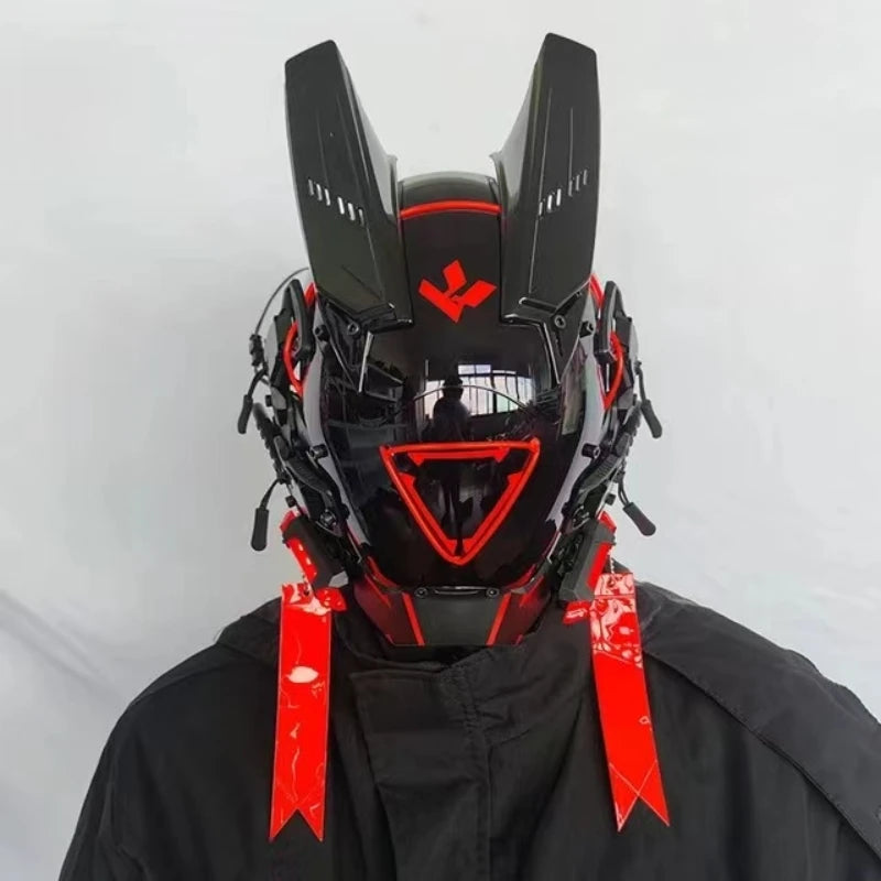 A person in black attire wears a futuristic HeadphoneHeat LED Lights Cyberpunk Full Face Mask. It features red and black accents, angular antenna-like extensions, a reflective visor, intricate side details, and LED lights on top for a sleek robotic look.