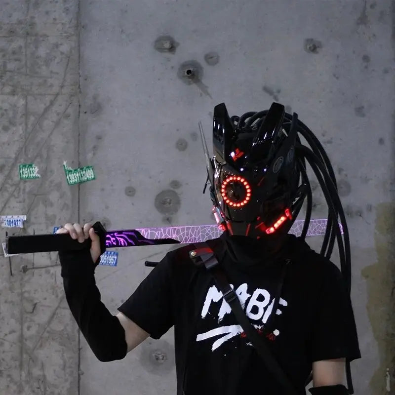 Wearing a HeadphoneHeat LED Lights Cyberpunk Full Face Mask with glowing red lights, a person holds a sword while donning a black outfit featuring graphic designs. Behind them, numbers and stickers adorn the concrete wall.