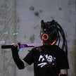 Wearing a HeadphoneHeat LED Lights Cyberpunk Full Face Mask with glowing red lights, a person holds a sword while donning a black outfit featuring graphic designs. Behind them, numbers and stickers adorn the concrete wall.