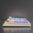 A side view of the HeadphoneHeat K82 Mechanical Gaming Keyboard showcases its colorful keycaps, featuring a gradient of blue, white, and orange. The backlighting on this wired mechanical keyboard radiates in shades of pink and blue, creating a vibrant effect against the dark background.