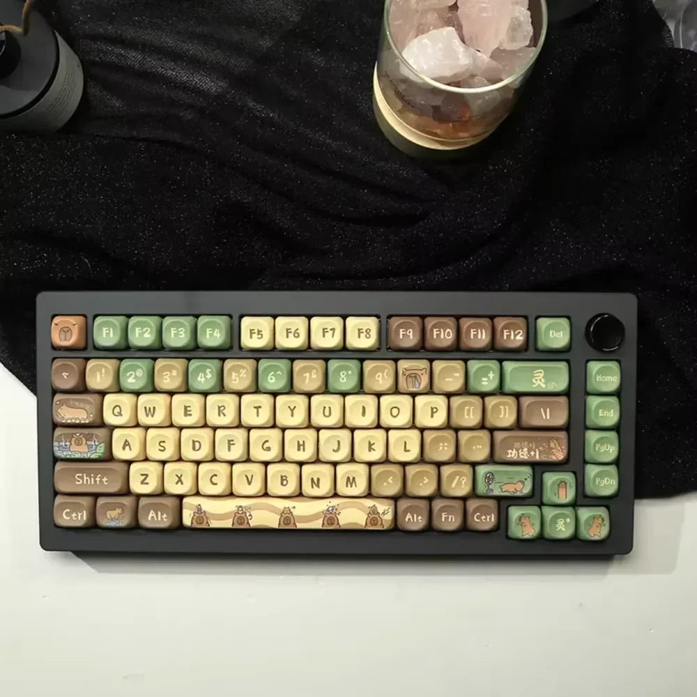 Introducing the HeadphoneHeat Capi Bara Mechanical Keyboard, featuring green and brown keys with nature-themed motifs like tree rings and leaves. This ergonomic design complements a black cloth setting with a glass of ice cubes, offering convenient seamless wireless technology.