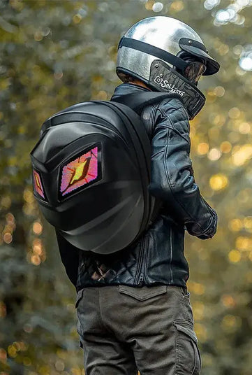 A person radiating an adventurous moto & biker style is walking outdoors, wearing a leather jacket and helmet, and carrying the HeadphoneHeat LED Turtle Shell Backpack. The lush greenery in the softly blurred background enhances the adventure-ready aura of this ABS material backpack.