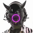 A person wears a HeadphoneHeat LED Lights Cyberpunk Full Face Mask featuring an LED-lit circle on the faceplate and horn-like top structures. The glossy black helmet boasts intricate mechanical details, perfectly complementing a striking black and white patterned outfit.