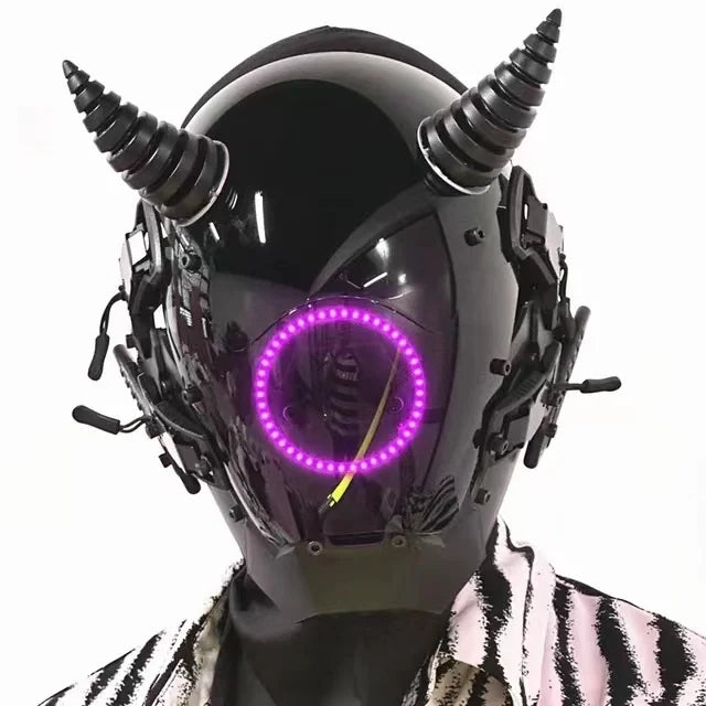 A person wears a HeadphoneHeat LED Lights Cyberpunk Full Face Mask featuring an LED-lit circle on the faceplate and horn-like top structures. The glossy black helmet boasts intricate mechanical details, perfectly complementing a striking black and white patterned outfit.