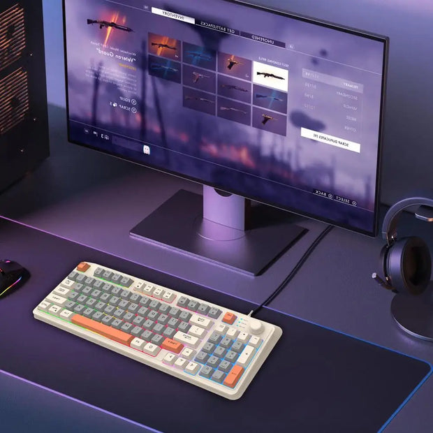 A computer monitor displays a video game interface with weapons selection. Below the monitor, a colorful K82 Mechanical Gaming Keyboard sits on a desk with a large mouse pad. A HeadphoneHeat headset rests on a stand nearby, all illuminated by soft, ambient lighting.