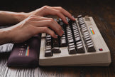 Hands typing on the 8BitDo Retro Mechanical Keyboard by HeadphoneHeat, featuring gray keys and a beige body. A wrist rest rests in front on the dark wood table. This vintage piece, adorned with small, colorful stripes in the top right corner, evokes nostalgia reminiscent of an 8Bitdo accessory.