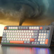 In front of a swirling tornado in the cloudy sky, a K82 Mechanical Gaming Keyboard by HeadphoneHeat with colorful keycaps and a USB interface sits dramatically. This wired mechanical keyboard features a convenient volume knob on the right side.