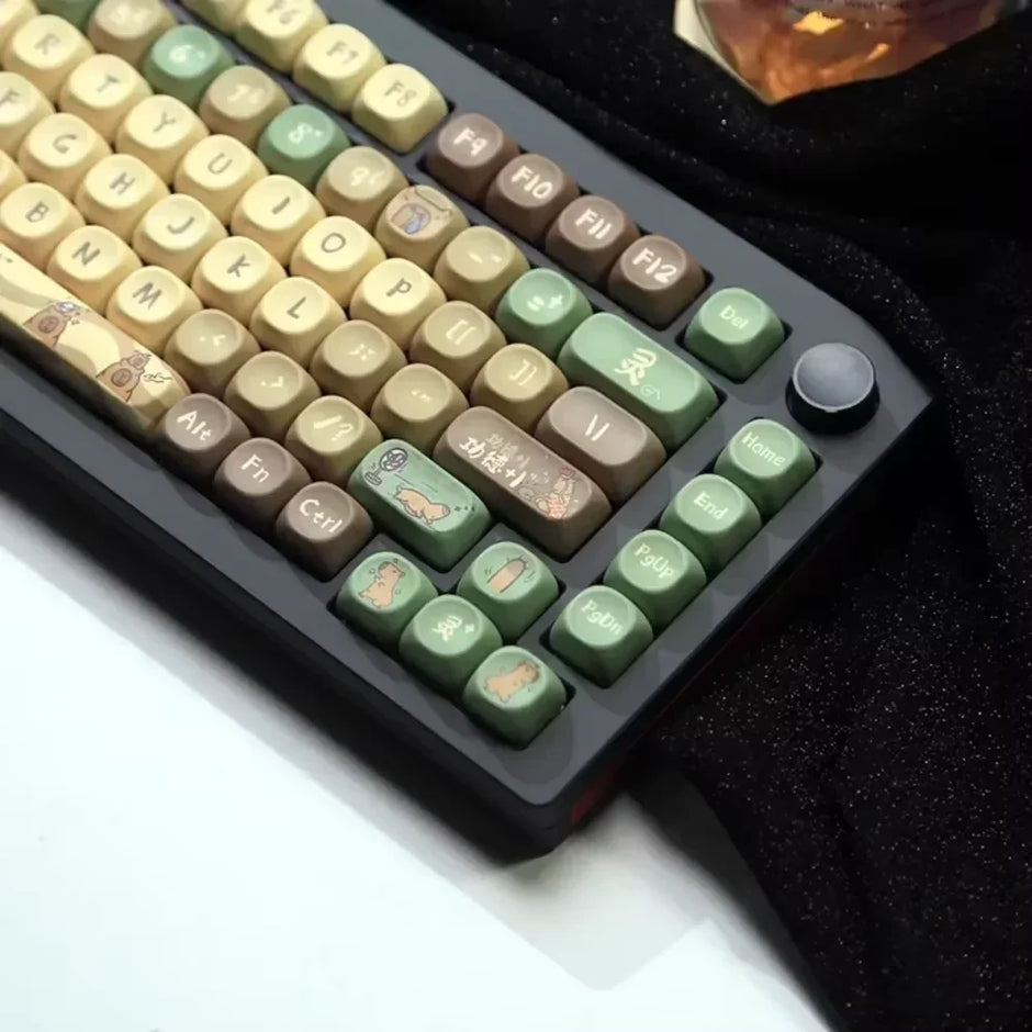The Capi Bara Mechanical Keyboard by HeadphoneHeat features unique keycaps with cartoon characters and symbols. Its ergonomic design offers comfort in green, brown, and beige. The wireless tech is enhanced by a black knob and function keys on a dark cloth surface.