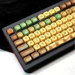 Introducing the Capi BaraCapi Bara Mechanical Key Keyboard by HeadphoneHeat—a wireless mechanical keyboard featuring ergonomically designed round keycaps in green, yellow, and brown hues. Each keycap displays unique retro-style icons such as a house and a tree, while the entire keyboard rests elegantly on a sleek black surface.