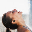 A woman wearing FLIPSTYLEZLLC Subwoofer Class 7 Waterproof True Wireless Earbuds enjoys crisp stereo sound and refreshment under a beach shower, smiling as water runs over her Bluetooth in-ear TWS headphones.