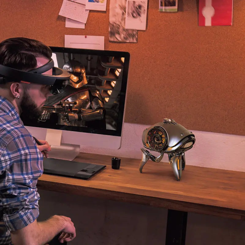 A man wearing augmented reality glasses admires a futuristic clock on his desk beside the Dropshipman Alien Tripod Bluetooth Speaker, while a desktop computer shows a robot scene and a corkboard decorates the organized workspace wall.