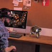 A man wearing augmented reality glasses admires a futuristic clock on his desk beside the Dropshipman Alien Tripod Bluetooth Speaker, while a desktop computer shows a robot scene and a corkboard decorates the organized workspace wall.