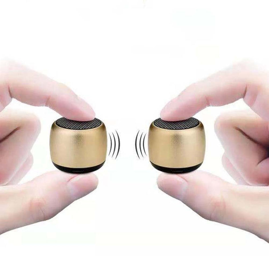 Two fingers on each side hold small, gold-colored Portable Mini Wireless Bluetooth Speakers from HeadphoneHeat.Com, facing each other. Sound wave symbols show enhanced sound quality against a plain white backdrop, highlighting their compact and durable design.
