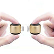 Two fingers on each side hold small, gold-colored Portable Mini Wireless Bluetooth Speakers from HeadphoneHeat.Com, facing each other. Sound wave symbols show enhanced sound quality against a plain white backdrop, highlighting their compact and durable design.